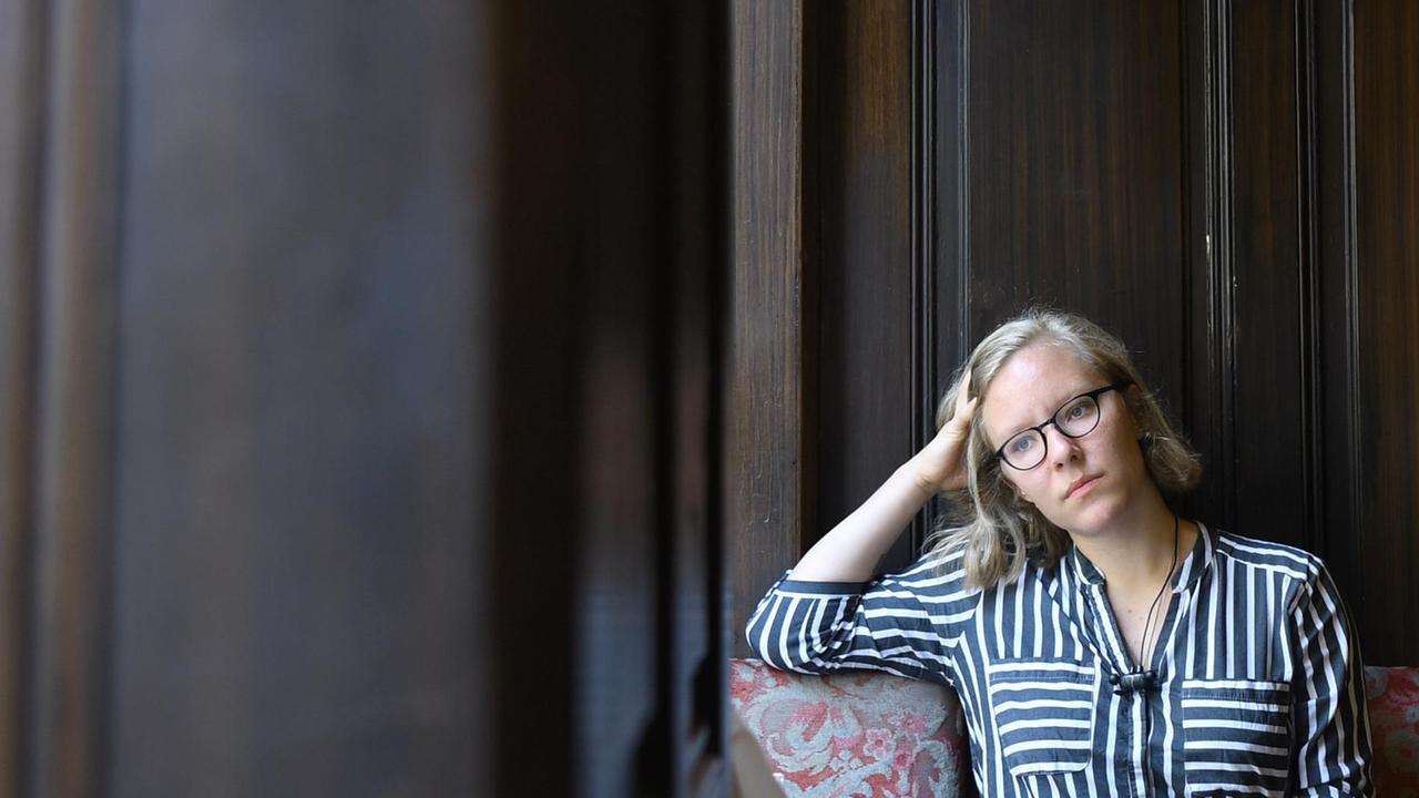 Das Bild zeigt die die österreichische Schriftstellerin Raphaela Edelbauer, die vor einer dunklen Holzwand sitzt. Das Bild zeigt die die österreichische Schriftstellerin Raphaela Edelbauer, die vor einer dunklen Holzwand sitzt.