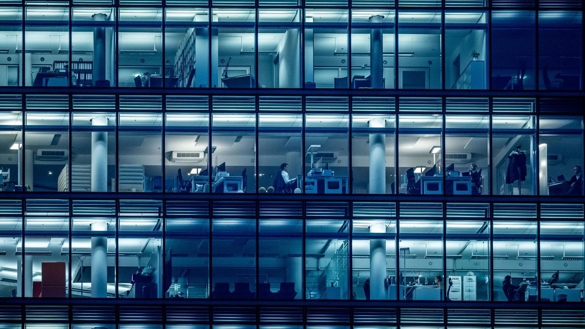 In einem Bürogebäude sind durch die Fenster einzelne Menschen zu sehen, die an Schreibtischen sitzen. In einem Bürogebäude sind durch die Fenster einzelne Menschen zu sehen, die an Schreibtischen sitzen.