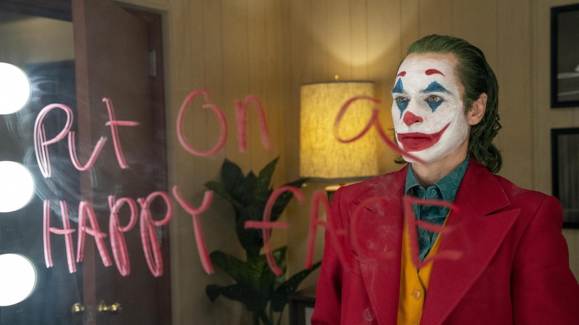 Joaquin Phoenix als Joker steht vor einem Spiegel, wodrauf in roter Schrift steht "Put on a happy face"
