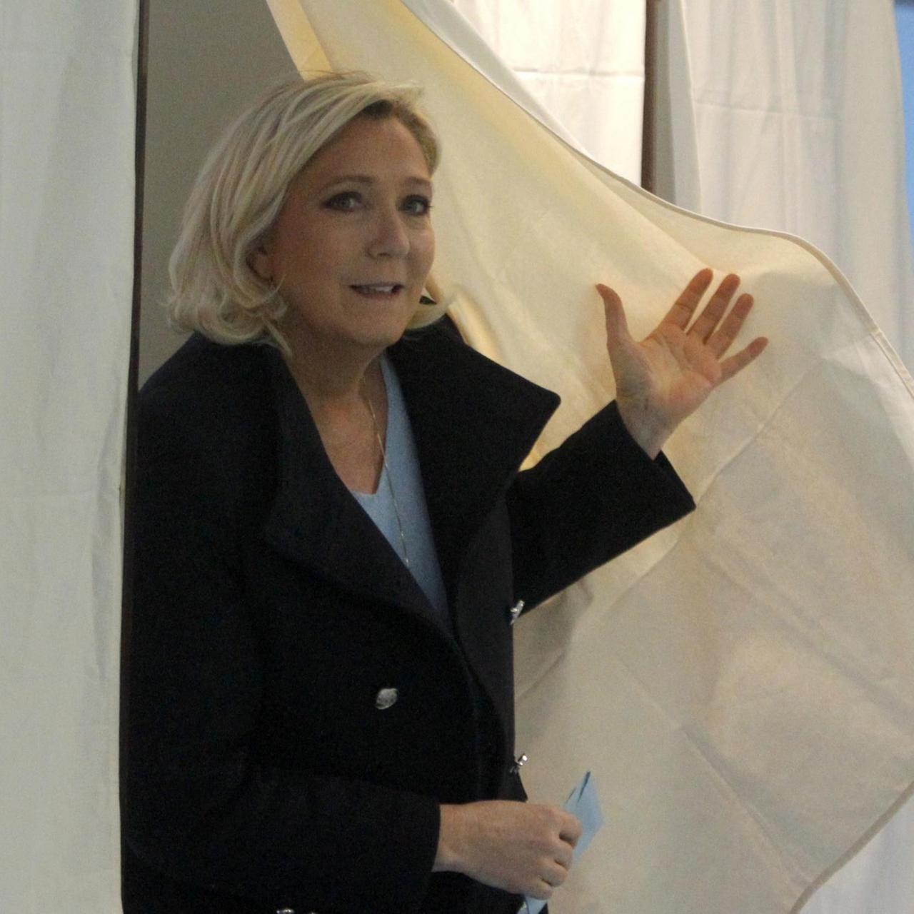 Marine Le pen kommt aus der Wahlkabine zurück. Sie schiebt mit ihrer linken Hand den Vorhang auf Seite und hält in ihrer rechten Hand den Umschlag mit dem Stimmzettel. 