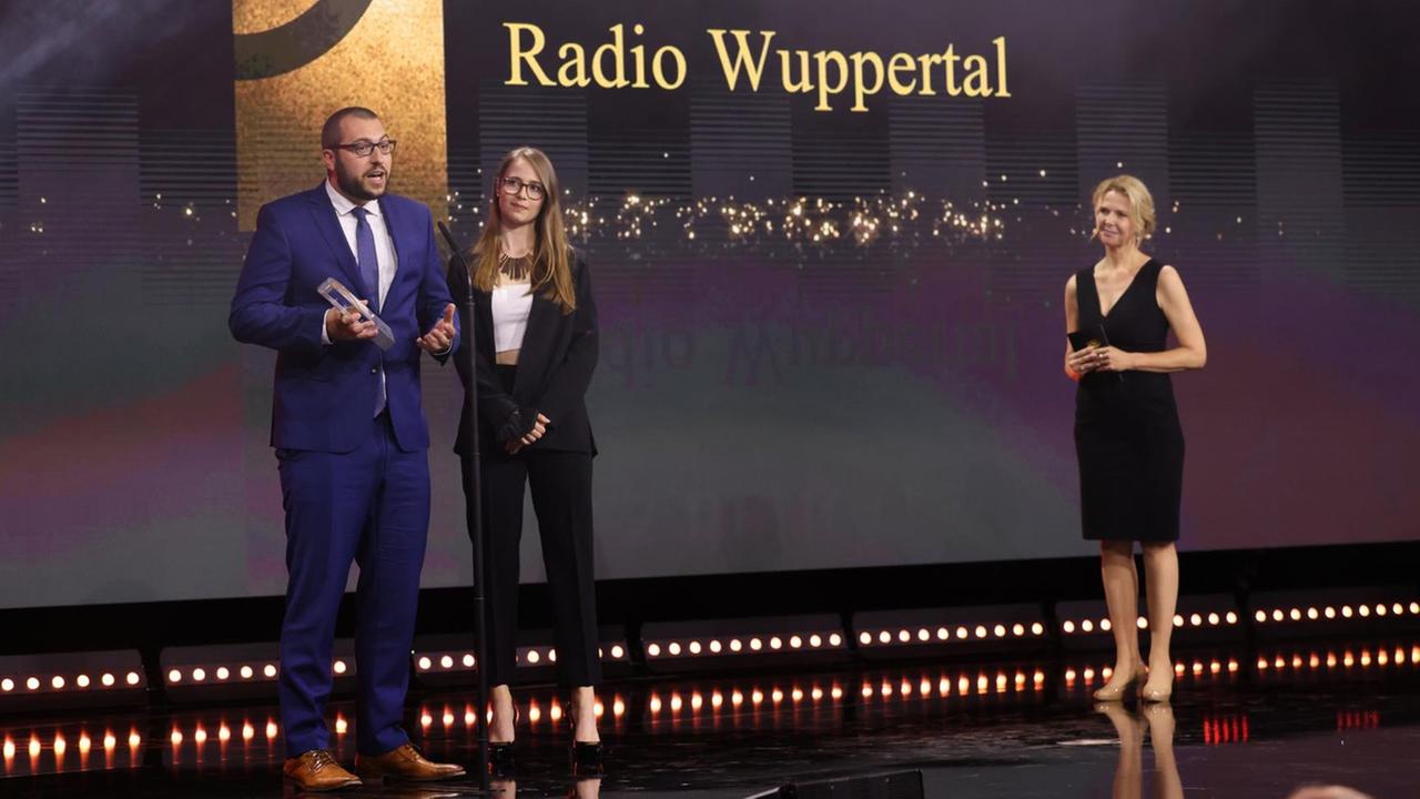 Jens Voss und Laura Mertens von Radio Wuppertal stehen bei der Gala des Deutschen Radiopreises 2021 mit ihrer Auszeichnung an einem Mikrophon. Jens Voss und Laura Mertens von Radio Wuppertal stehen bei der Gala des Deutschen Radiopreises 2021 mit ihrer Auszeichnung an einem Mikrophon.