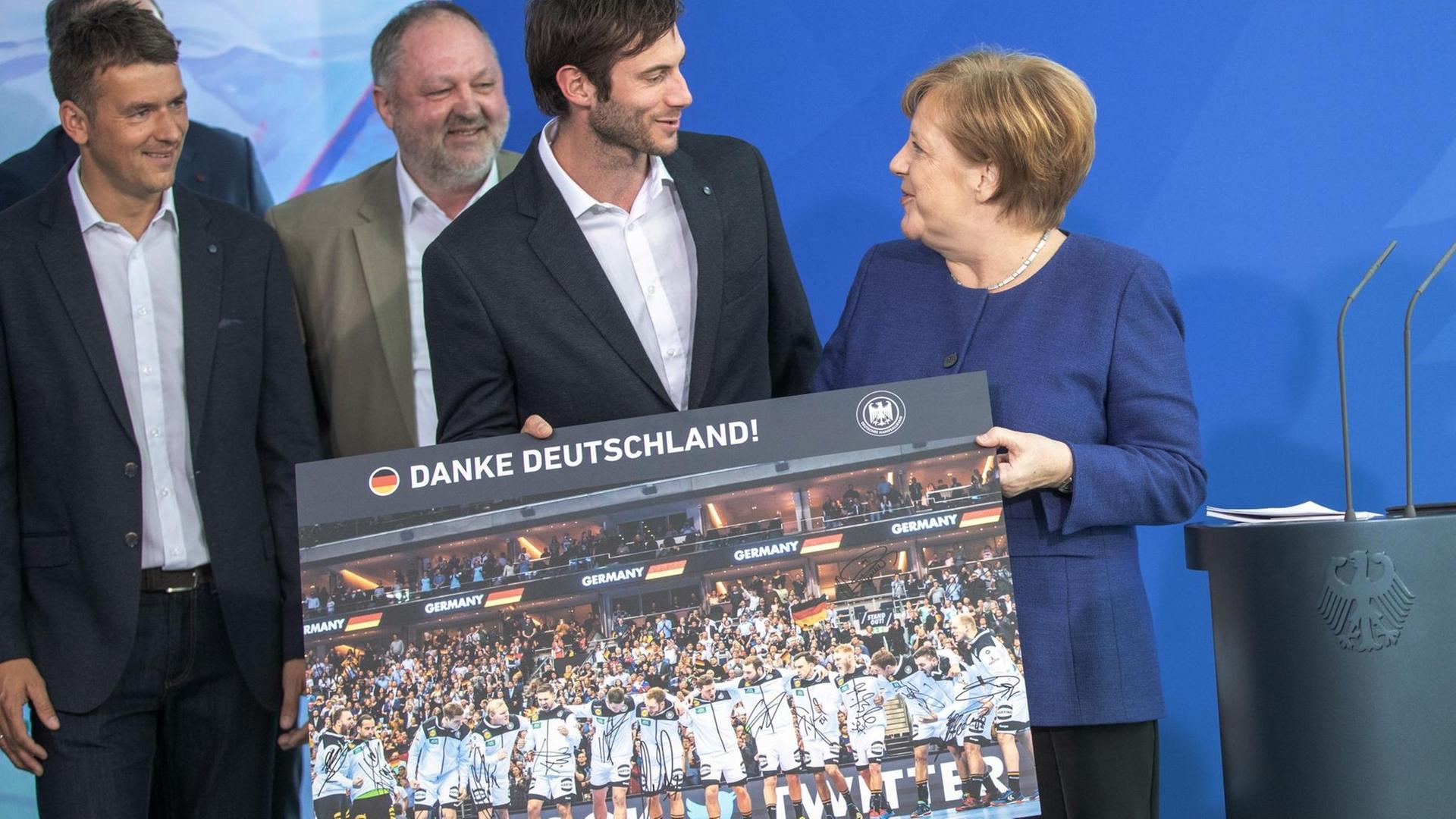 Bundeskanzlerin Angela Merkel (CDU) nimmt beim Empfang für die deutsche Handball-Nationalmannschaft im Bundeskanzleramt ein Poster von Teamkapitän Uwe Gensheimer entgegen. im Hintergrund Bundestrainer Christian Prokop (l) und Handball-Präsident Andreas Michelmann.