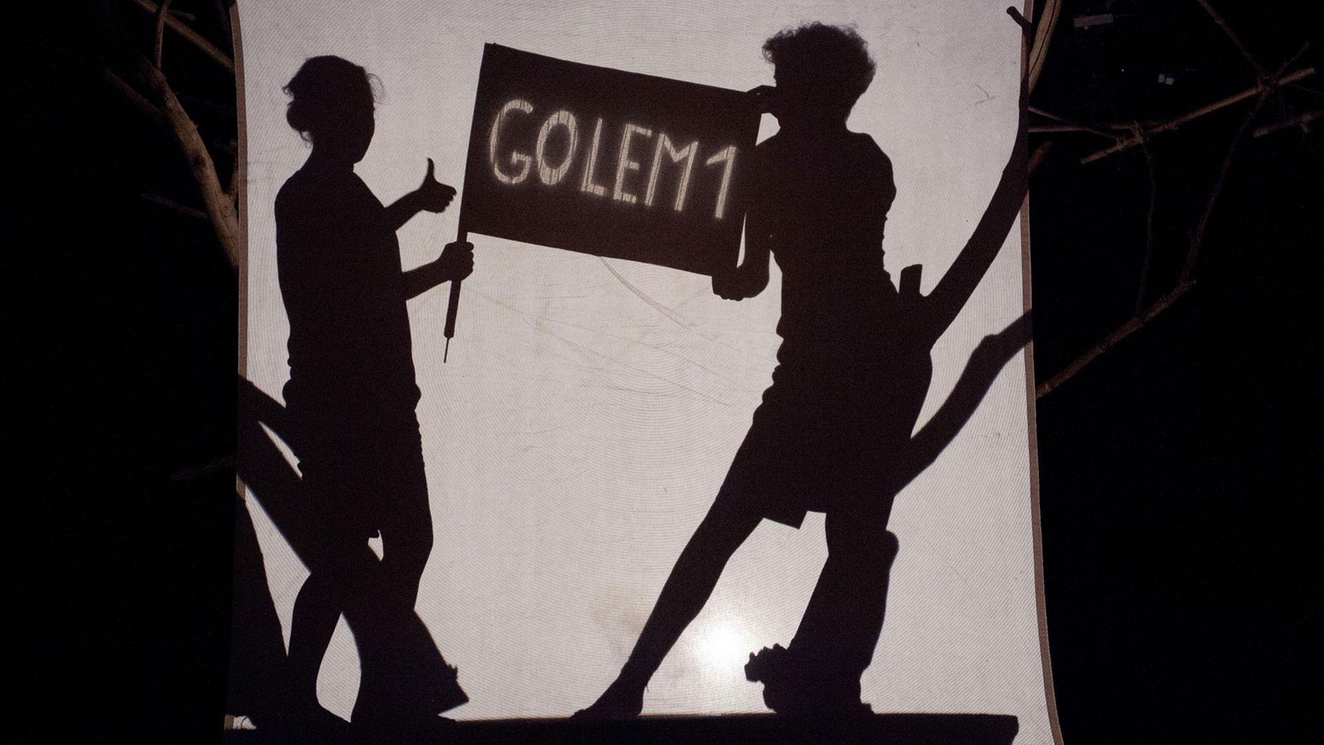 Szene aus "Golem": Eine Frau und ein Mann halten ein Schild mit der Aufschrift "Golem1" - alles ist als Schattenriss zu sehen. Szene aus "Golem": Eine Frau und ein Mann halten ein Schild mit der Aufschrift "Golem1" - alles ist als Schattenriss zu sehen.