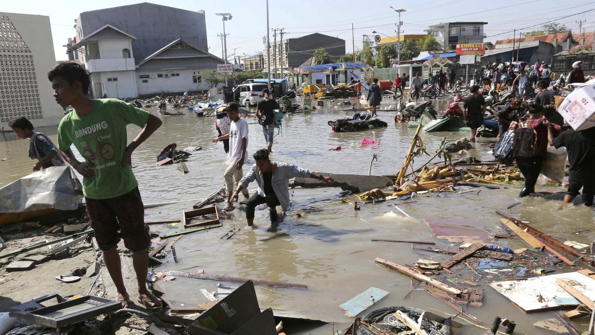 Die Menschen waten durch ein überflutetes Gelände in der Stadt; im Wasser schwimmen Trümmer.