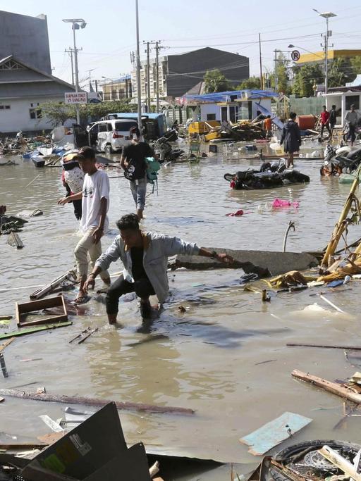 Die Menschen waten durch ein überflutetes Gelände in der Stadt; im Wasser schwimmen Trümmer.