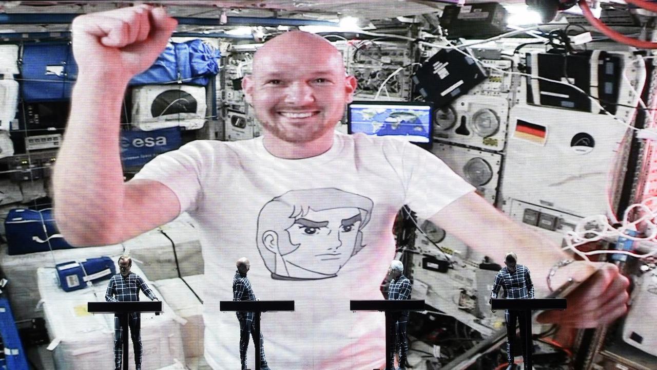 Die Band Kraftwerk auf der Bühne. Dahinter in der Videoschalte: Der Astronaut Alexander Gerst mit einem Tablet-Computer. Er macht eine triumphierende Geste. Die Band Kraftwerk auf der Bühne. Dahinter in der Videoschalte: Der Astronaut Alexander Gerst mit einem Tablet-Computer. Er macht eine triumphierende Geste.