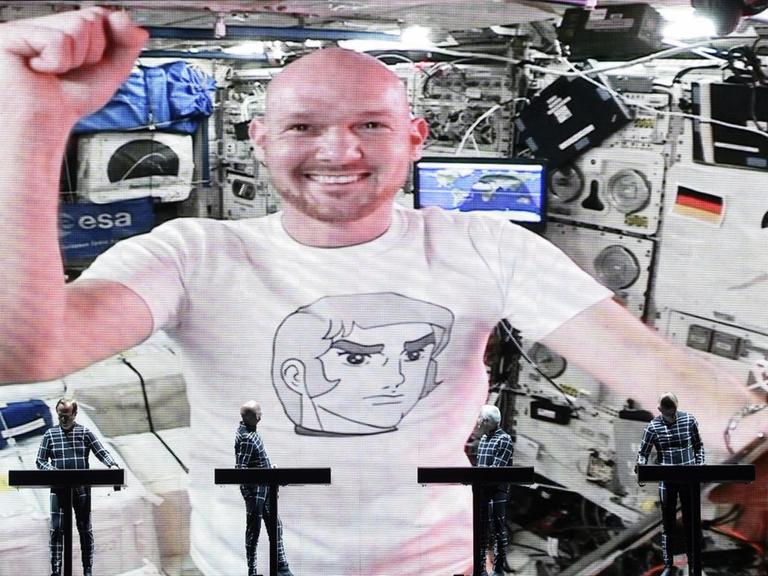 Die Band Kraftwerk auf der Bühne. Dahinter in der Videoschalte: Der Astronaut Alexander Gerst mit einem Tablet-Computer. Er macht eine triumphierende Geste.