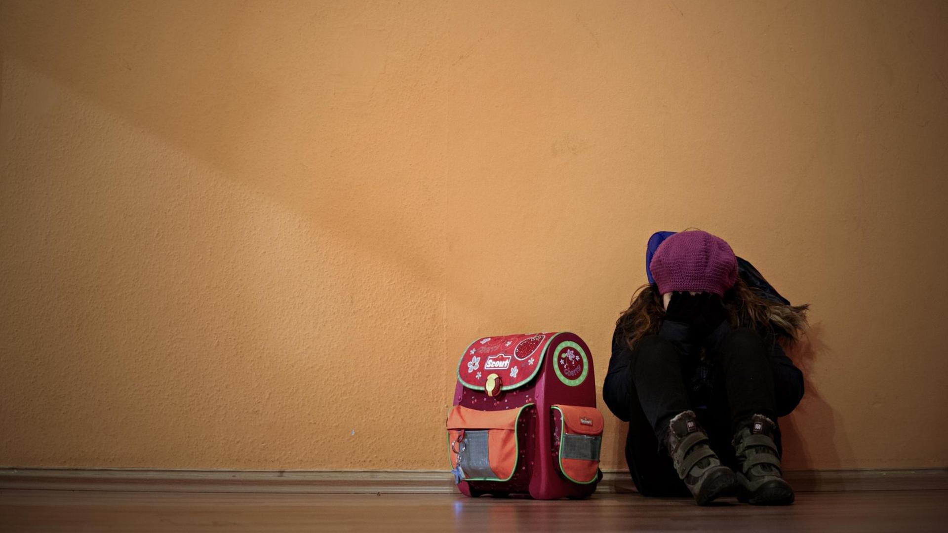 Ein kleines Mädchen sitzt an eine Wand gelehnt neben ihrem Schulranzen und hält sich die Hände vors Gesicht. Ein kleines Mädchen sitzt an eine Wand gelehnt neben ihrem Schulranzen und hält sich die Hände vors Gesicht.
