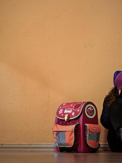 Ein kleines Mädchen sitzt an eine Wand gelehnt neben ihrem Schulranzen und hält sich die Hände vors Gesicht.