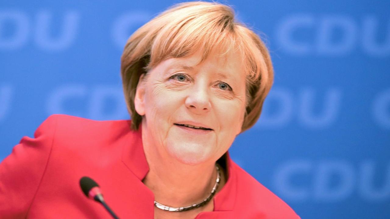 Merkel lächelnd im roten Kostüm vor einem Mikrofon, dahinter eine blaue Wand mit "CDU-Schriftzügen". Merkel lächelnd im roten Kostüm vor einem Mikrofon, dahinter eine blaue Wand mit "CDU-Schriftzügen".