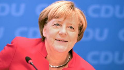 Merkel lächelnd im roten Kostüm vor einem Mikrofon, dahinter eine blaue Wand mit "CDU-Schriftzügen".