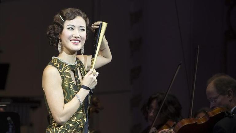 Eine junge Frau in grünem Kleid und Fächer steht vor einem Orchester und lächelt.