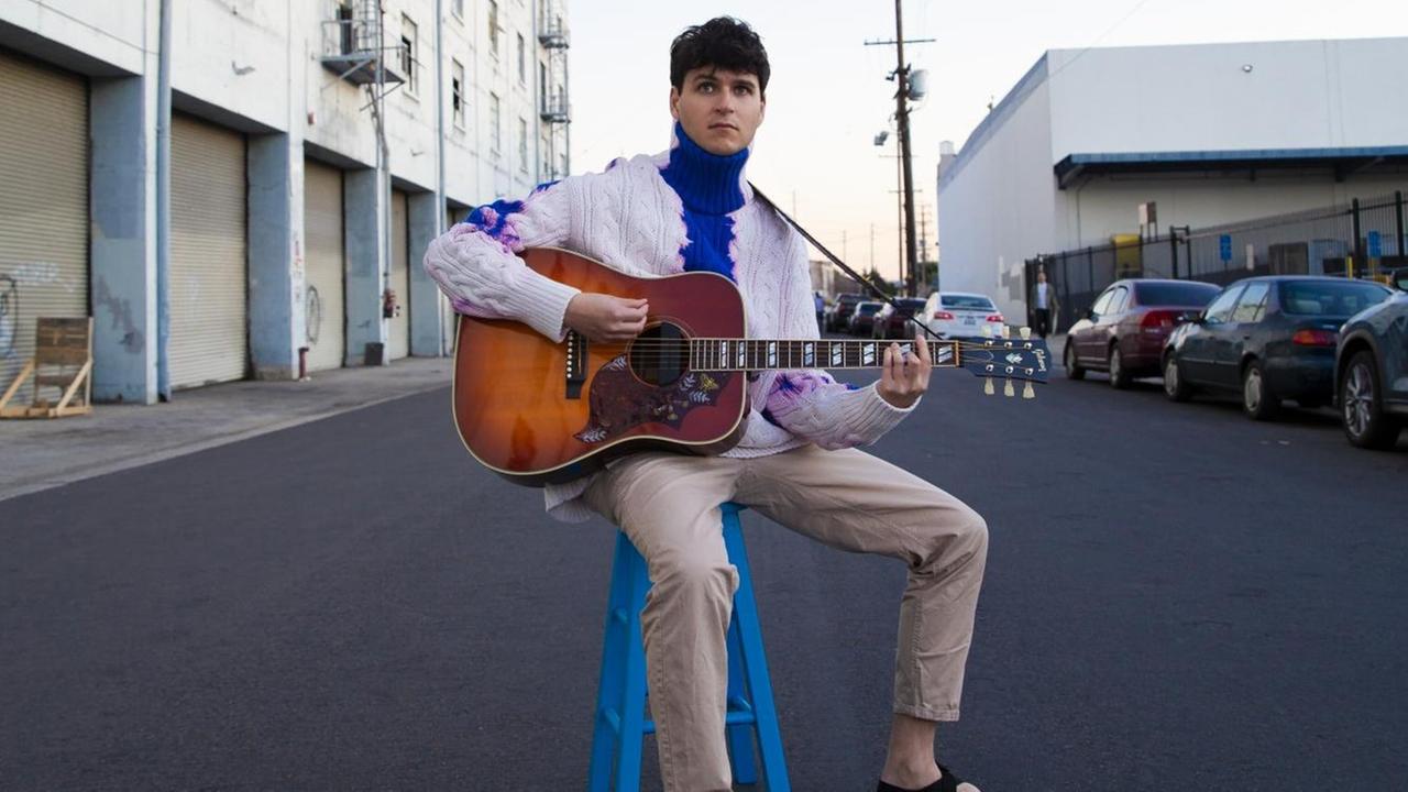 Der Frontmann von Vampire Weekend, Ezra Koenig, spielt mitten auf einer Straße auf einem blauen Hocker Gitarre