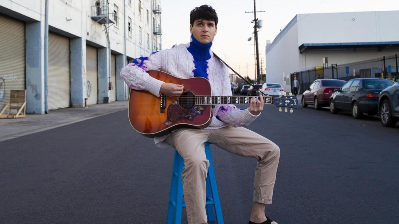 Der Frontmann von Vampire Weekend, Ezra Koenig, spielt mitten auf einer Straße auf einem blauen Hocker Gitarre Der Frontmann von Vampire Weekend, Ezra Koenig, spielt mitten auf einer Straße auf einem blauen Hocker Gitarre