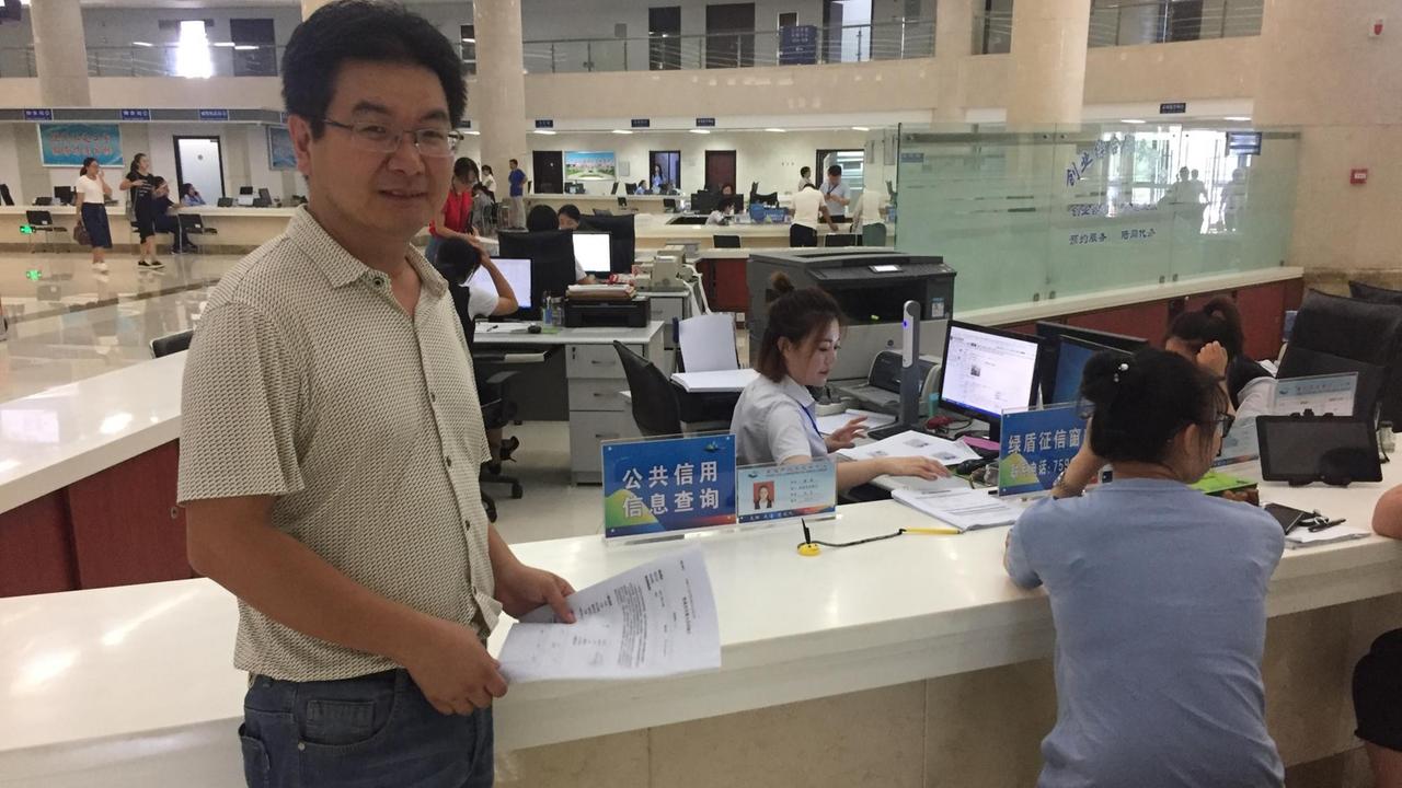Der Forstamt-Mitarbeiter Zhang Jian trägt ein weißes Hemd und steht im Bürgeramt, in dem viele Frauen an Computern sitzen. Der Forstamt-Mitarbeiter Zhang Jian trägt ein weißes Hemd und steht im Bürgeramt, in dem viele Frauen an Computern sitzen.