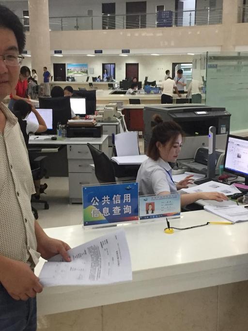 Der Forstamt-Mitarbeiter Zhang Jian trägt ein weißes Hemd und steht im Bürgeramt, in dem viele Frauen an Computern sitzen.