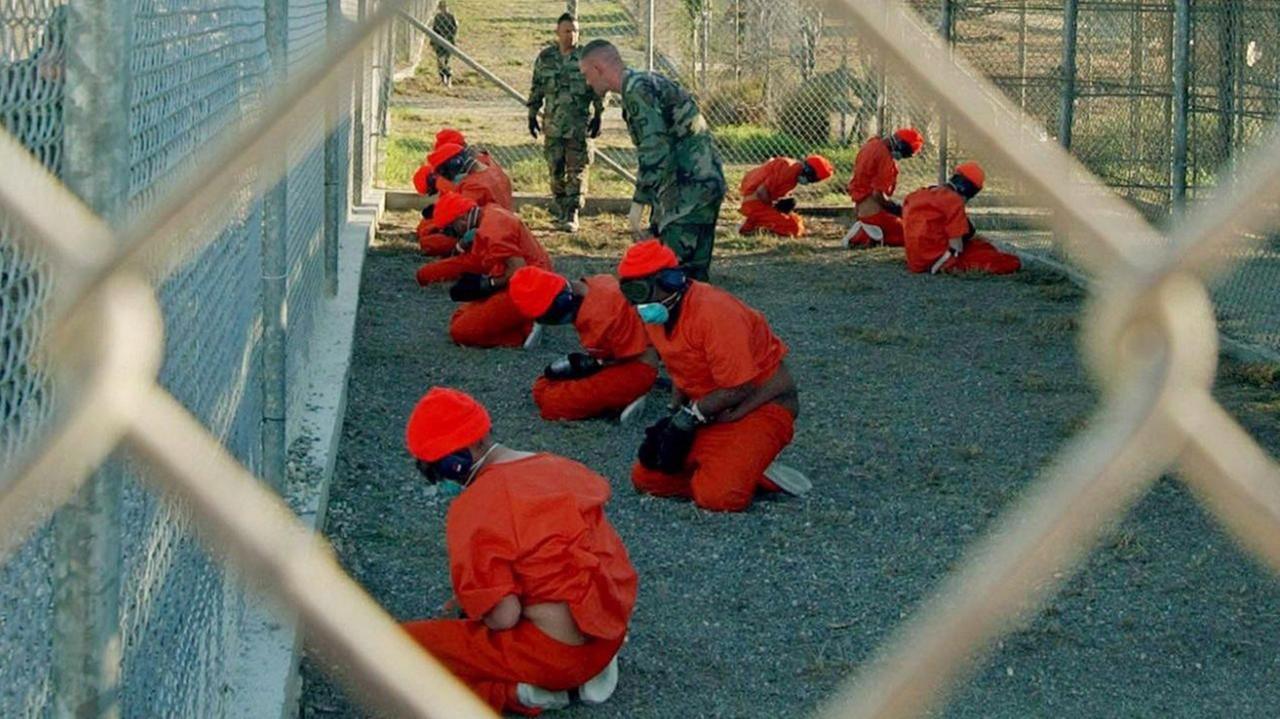 Häftlinge im Gefängnis Guantanamo im Jahr 2002. Sie sitzen auf dem Boden in roten Anzügen hinter einem Drahtzaun.