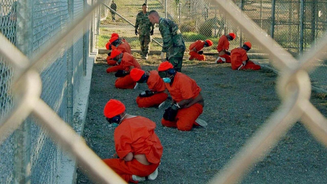 Häftlinge im Gefängnis Guantanamo im Jahr 2002. Sie sitzen auf dem Boden in roten Anzügen hinter einem Drahtzaun. Häftlinge im Gefängnis Guantanamo im Jahr 2002. Sie sitzen auf dem Boden in roten Anzügen hinter einem Drahtzaun.