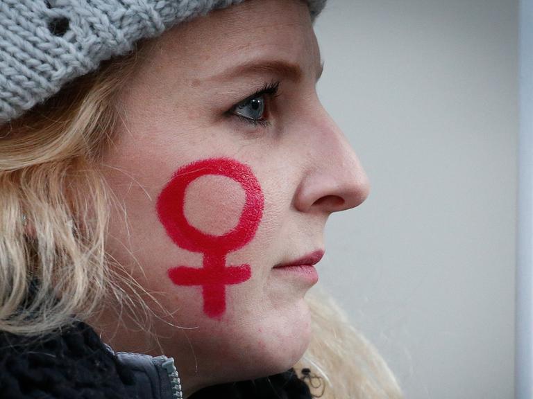 Das Bild zeigt das Foto einer jungen Frau, auf deren Wange mit rotem Stift das weibliche Gender-Symbol gezeichnet ist.