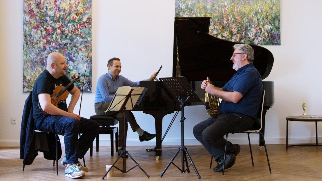 Die drei Musiker sitzen bei einer Probe in einem hohen Raum mit abstrakten Gemälden und lachen einander zu.