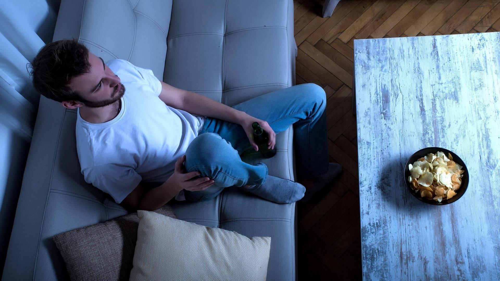 Draufsicht auf einen jungen Mann, der entspannt auf einer Couch sitzt und vor sich einen Tisch stehen hat, auf dem sich eine Schale Chips befindet. Draufsicht auf einen jungen Mann, der entspannt auf einer Couch sitzt und vor sich einen Tisch stehen hat, auf dem sich eine Schale Chips befindet.