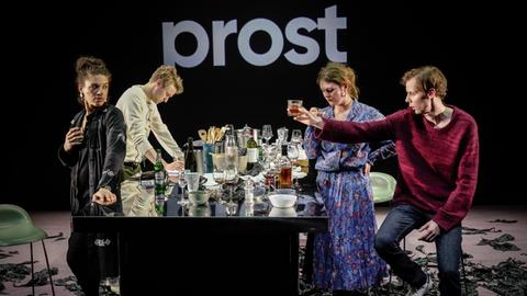 Vier Schauspieler an einem Tisch, auf dem viele leere Gläser und Flaschen stehen und prosten sich zu. Im Hintergrund steht weiß auf schwarzer Wand: "Prost".