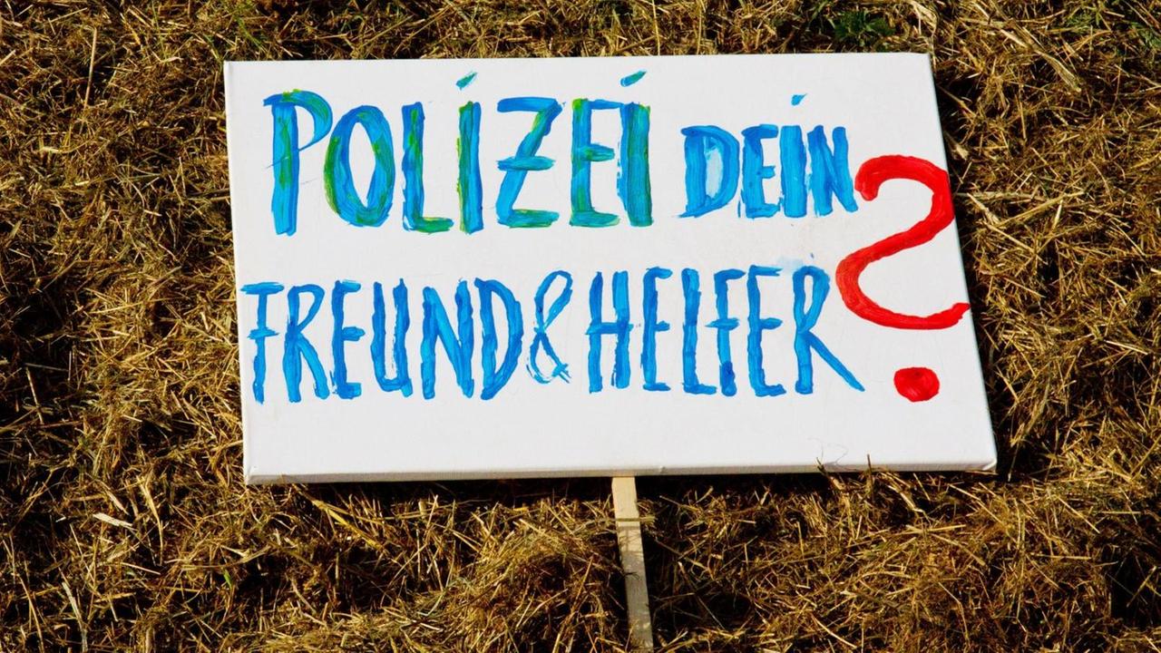"Polizei dein Freund und Helfer?", steht geschrieben auf einem Schild in blauen Buchstaben und mit rotem Fragenzeichen auf einer grünen Wiese liegend.