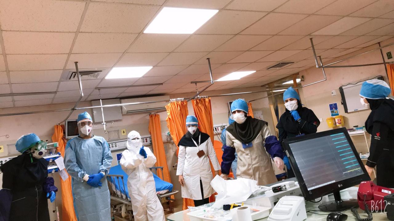 Ein Halbkreis von von Kopf bis Fuß vermummtem Plegepersonal, 7 Personen, steht um einen Tisch mit PC. Ein Halbkreis von von Kopf bis Fuß vermummtem Plegepersonal, 7 Personen, steht um einen Tisch mit PC.