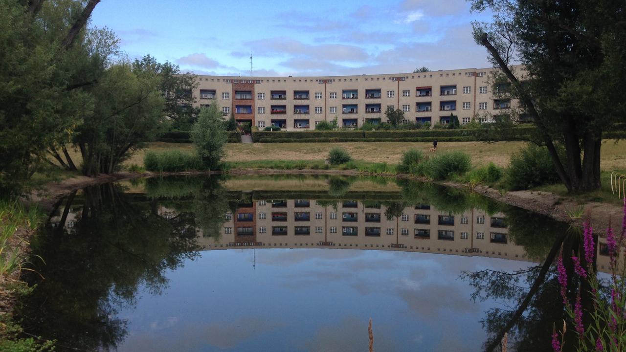 Das Bild zeigt hufeisenförmig angeordnete Häuser im Halbrund um einen See, der im Vordergrund ist. Die Hufeisensiedlung in Berlin-Britz. Das Bild zeigt hufeisenförmig angeordnete Häuser im Halbrund um einen See, der im Vordergrund ist. Die Hufeisensiedlung in Berlin-Britz.