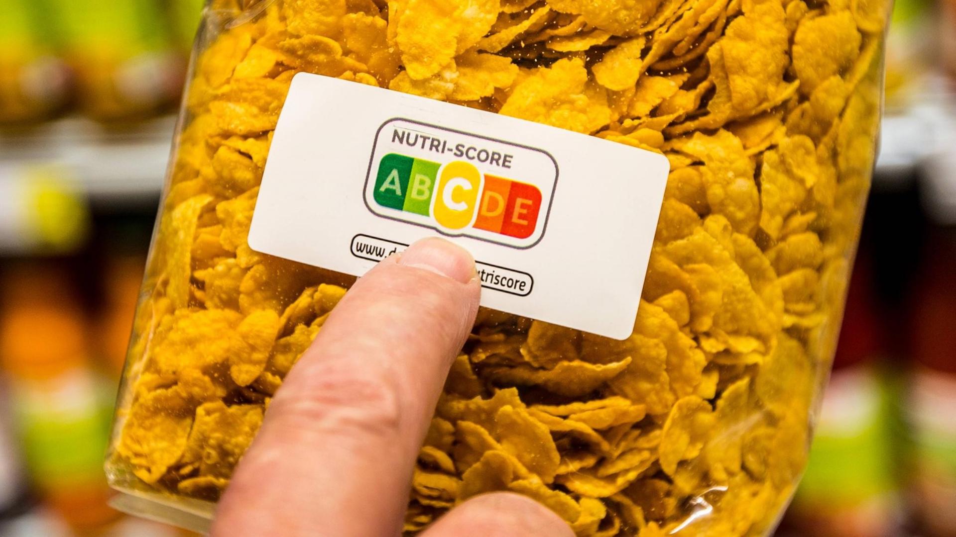Eine Packung Cornflakes, ein Zeigefinger deutet auf die Farbflächen des Nutri-Score-Aufdrucks. Die mittlere Kategorie "C" (gelb) ist hervorgehoben. Eine Packung Cornflakes, ein Zeigefinger deutet auf die Farbflächen des Nutri-Score-Aufdrucks. Die mittlere Kategorie "C" (gelb) ist hervorgehoben.