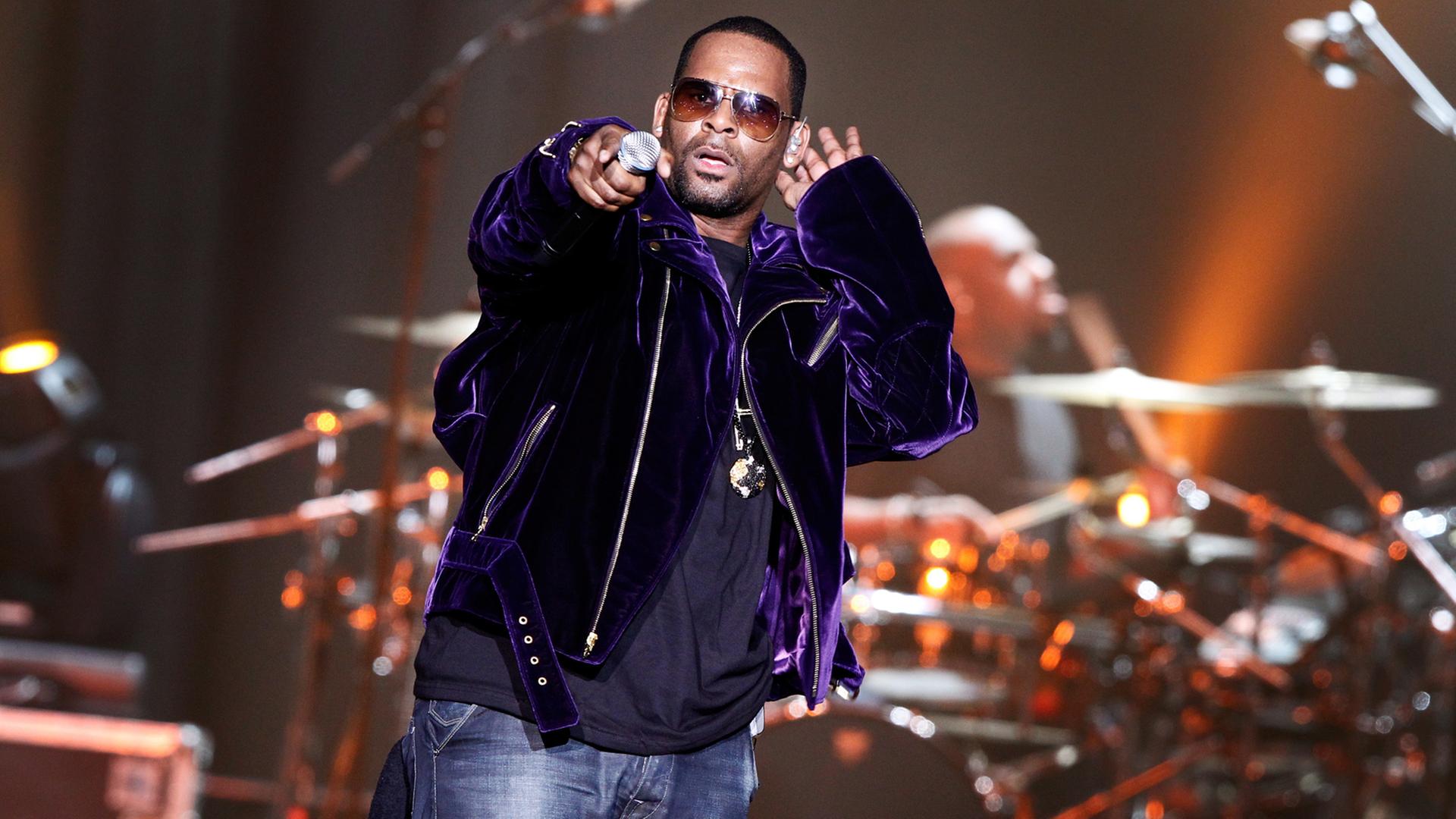 Der US-Sänger R. Kelly steht auf der Bühne, im Hintergrund ein Schlagzeug. Er hält sich eine Hand hinter sein linkes Ohr, mit der rechten Hand zeigt er ins Publikum.