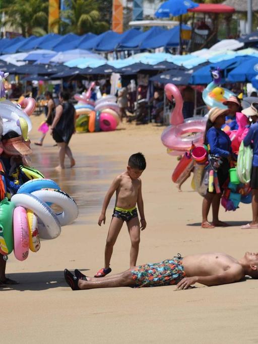 Strandszene in Acapulco mit Badegästen, Sonnenschirmen und Badetierverkäufern.