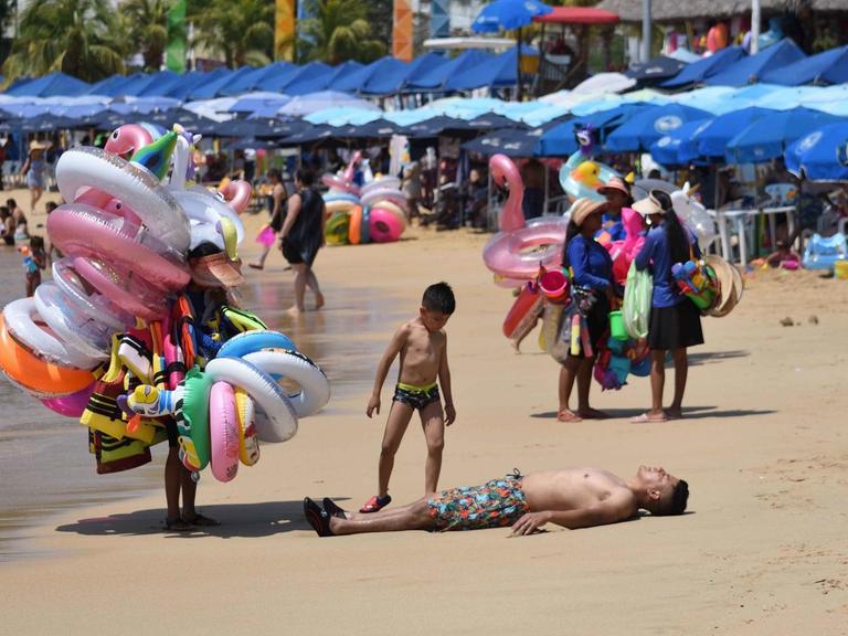 Strandszene in Acapulco mit Badegästen, Sonnenschirmen und Badetierverkäufern.
