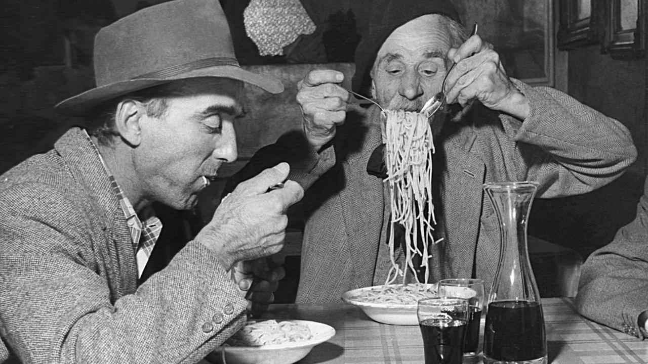Eine historische Fotografie von zwei Männern, die an einem Tisch Spagetti essen.