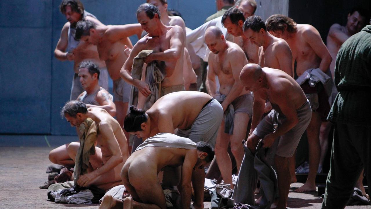 Viele Männer ent- oder bekleiden sich auf einer Bühne, einige sind komplett nackt, andere steigen gerade in Hosen oder ziehen T-Shirts oder Hemden an. Alle Stoffe sind in Grau-, Beige- und Weißtönen gehalten. Viele Männer ent- oder bekleiden sich auf einer Bühne, einige sind komplett nackt, andere steigen gerade in Hosen oder ziehen T-Shirts oder Hemden an. Alle Stoffe sind in Grau-, Beige- und Weißtönen gehalten.