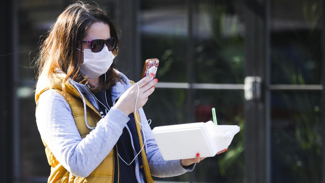 Eine Frau läuft durch eine Straße, trägt einen Mundschutz und Sonnenbrille und blickt dabei auf ihr Handy, das sie vor sich trägt. Eine Frau läuft durch eine Straße, trägt einen Mundschutz und Sonnenbrille und blickt dabei auf ihr Handy, das sie vor sich trägt.