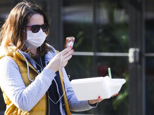 Eine Frau läuft durch eine Straße, trägt einen Mundschutz und Sonnenbrille und blickt dabei auf ihr Handy, das sie vor sich trägt.