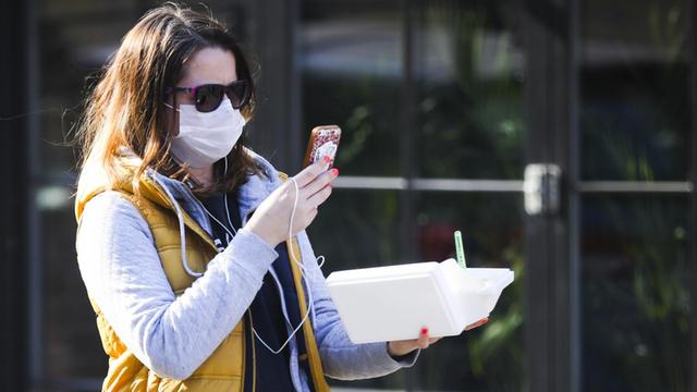Eine Frau läuft durch eine Straße, trägt einen Mundschutz und Sonnenbrille und blickt dabei auf ihr Handy, das sie vor sich trägt.