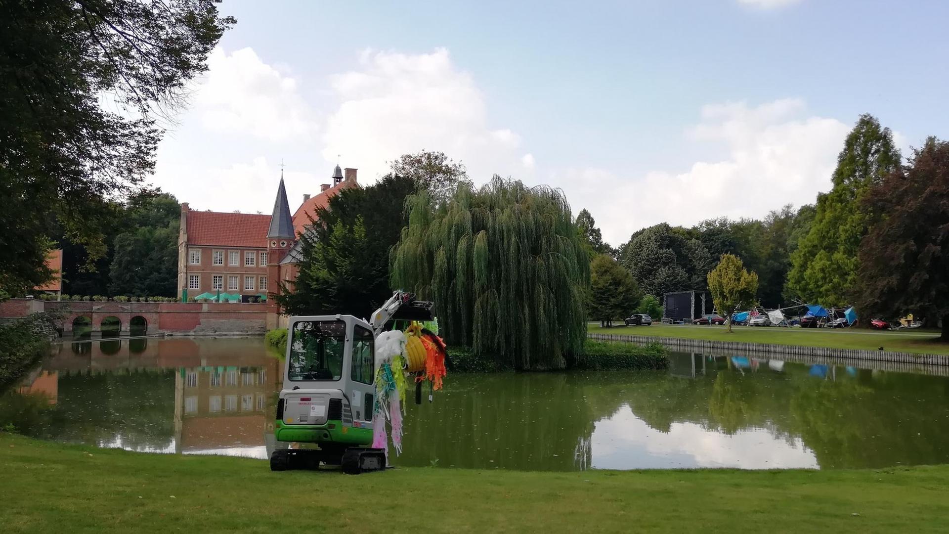 Im Hintergrund Burg Hülshoff, davor ein See, am Ufer ein dekorierter Bagger Im Hintergrund Burg Hülshoff, davor ein See, am Ufer ein dekorierter Bagger