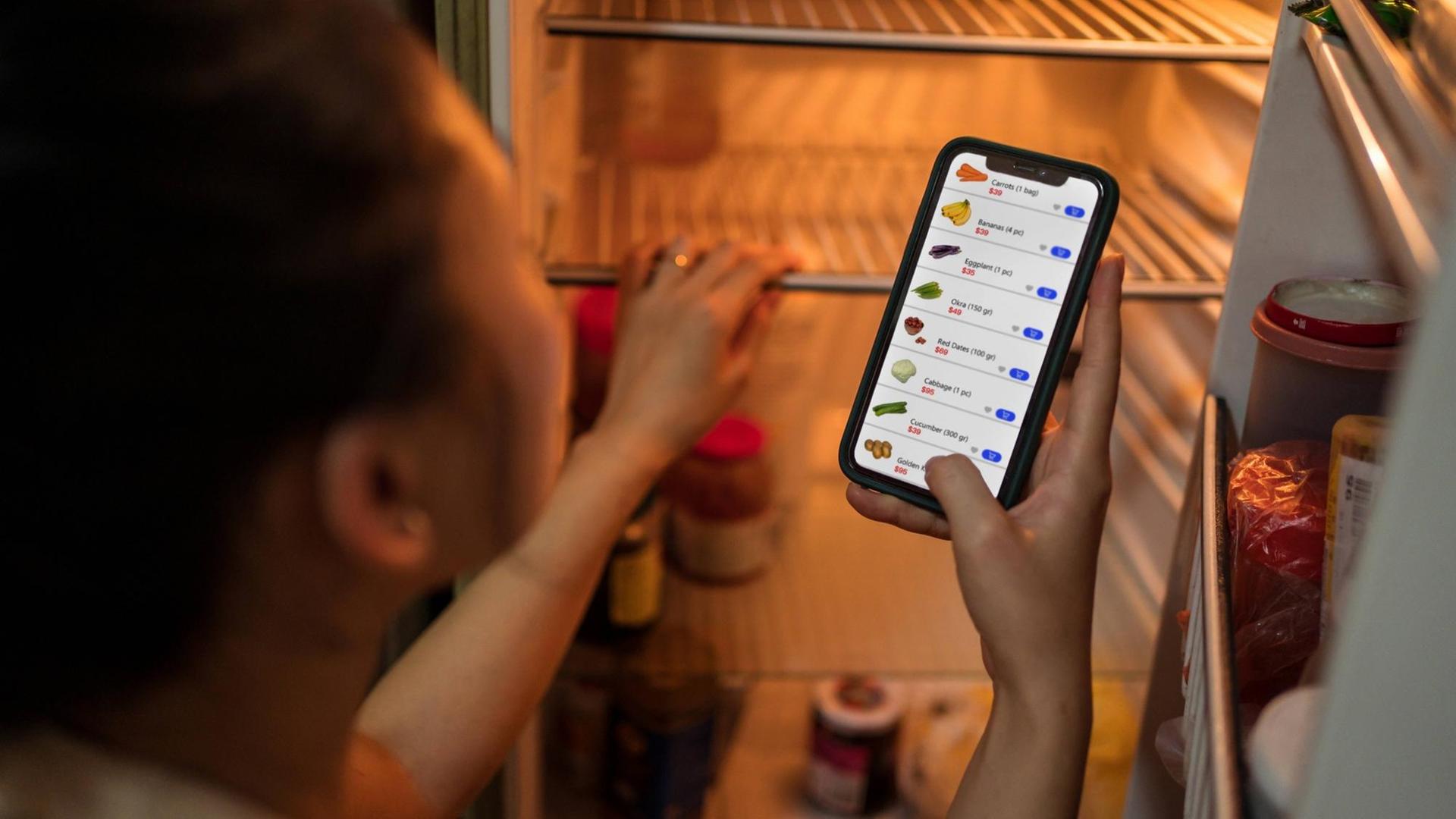 Eine Frau steht vor einem offenen Kühlschrank und bestellt Lebensmittel per App. Eine Frau steht vor einem offenen Kühlschrank und bestellt Lebensmittel per App.