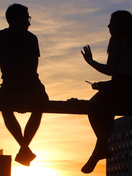 Zwei Personen sitzen auf dem Geländer der Hackerbrücke in München und genießen den Sonnenuntergang bei einer Unterhaltung.