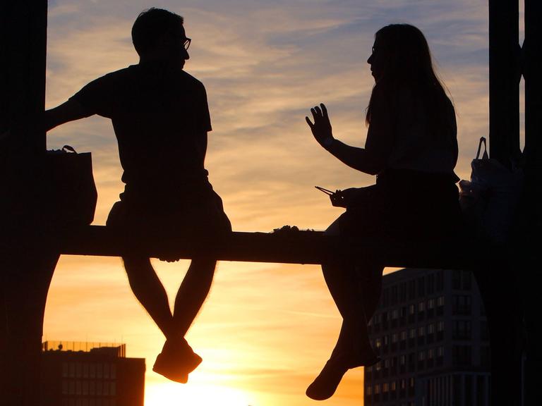 Zwei Personen sitzen auf dem Geländer der Hackerbrücke in München und genießen den Sonnenuntergang bei einer Unterhaltung.