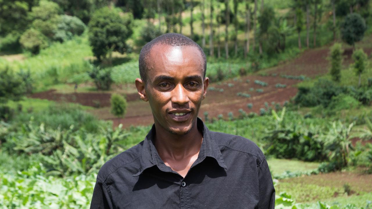 Ein schlanker, jüngerer afrikanischer Mann steht vor Bäumen und lächelt in die Kamera. Ein schlanker, jüngerer afrikanischer Mann steht vor Bäumen und lächelt in die Kamera.