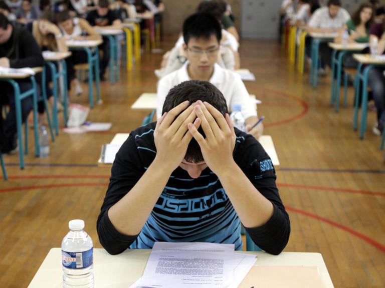 Schüler an Schreibtischen beim Pisa-Test. Der Schüler vorne im Bild hat seine Hände über dem Kopf gefaltet.
