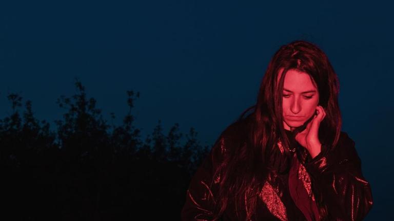 Lucia Cadotsch mit offenen, langen, dunklen Haaren wird rötlich auf einem Feld angestrahlt, während sie vor dunkelblauem Himmel steht - im Hintergrund zeichnet sich ein Wald ab. Lucia Cadotsch mit offenen, langen, dunklen Haaren wird rötlich auf einem Feld angestrahlt, während sie vor dunkelblauem Himmel steht - im Hintergrund zeichnet sich ein Wald ab.