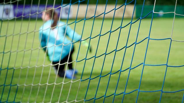 Foto aufgenommen durch das Netz eines Fußballtors: Torfrau in einem hellblauen Trikot kniet auf dem Rasen