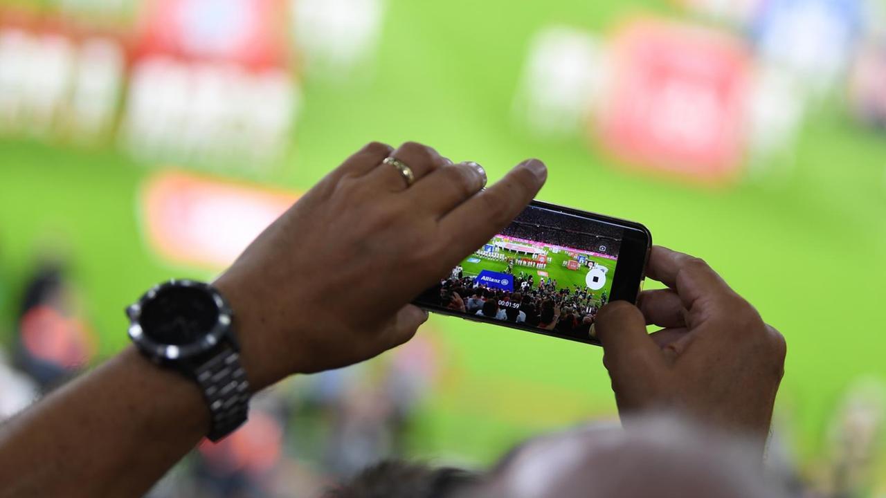 Stadionbesucher sieht das Geschehen auf dem Fußballplatz durch einen Smartphone-Bildschirm Stadionbesucher sieht das Geschehen auf dem Fußballplatz durch einen Smartphone-Bildschirm