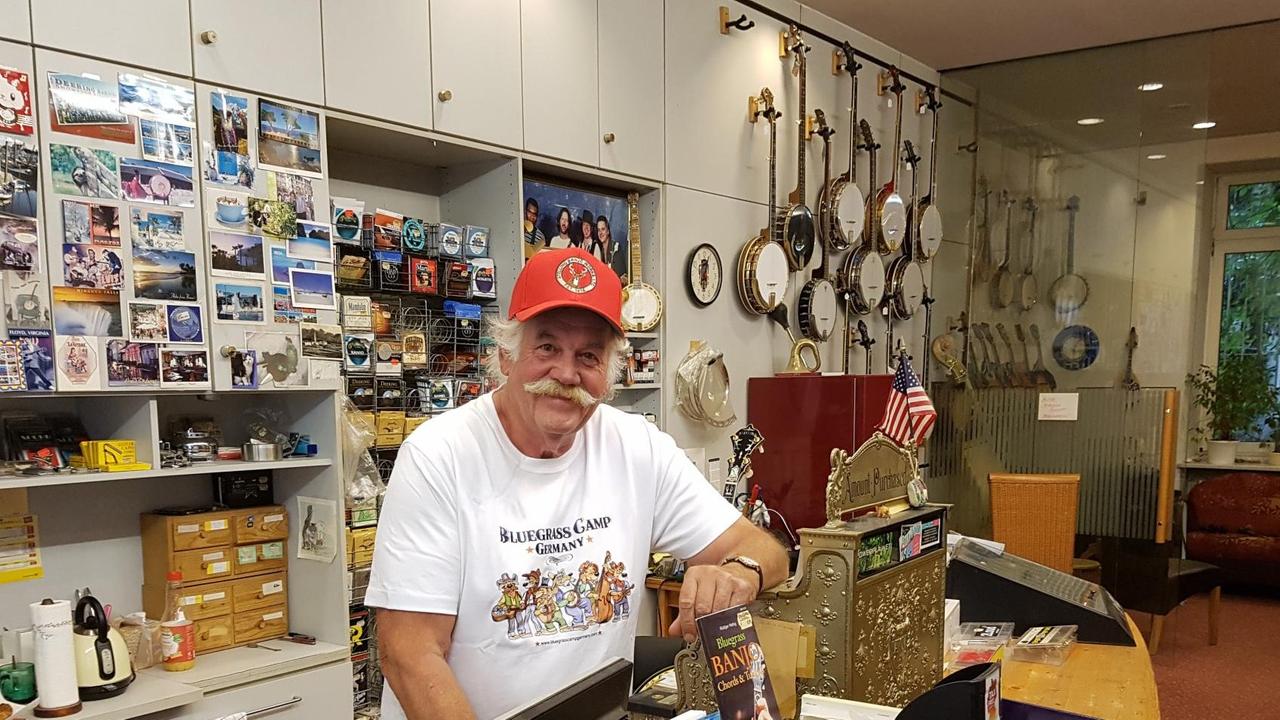 Ein Mann im weißen T-Shirt und roter Kappe lehnt sich an eine alte Registrierkasse. Er steht vor einer weißen Schrankwand. An der Schrankwand hängen Postkarten und Banjos. Ein Mann im weißen T-Shirt und roter Kappe lehnt sich an eine alte Registrierkasse. Er steht vor einer weißen Schrankwand. An der Schrankwand hängen Postkarten und Banjos.