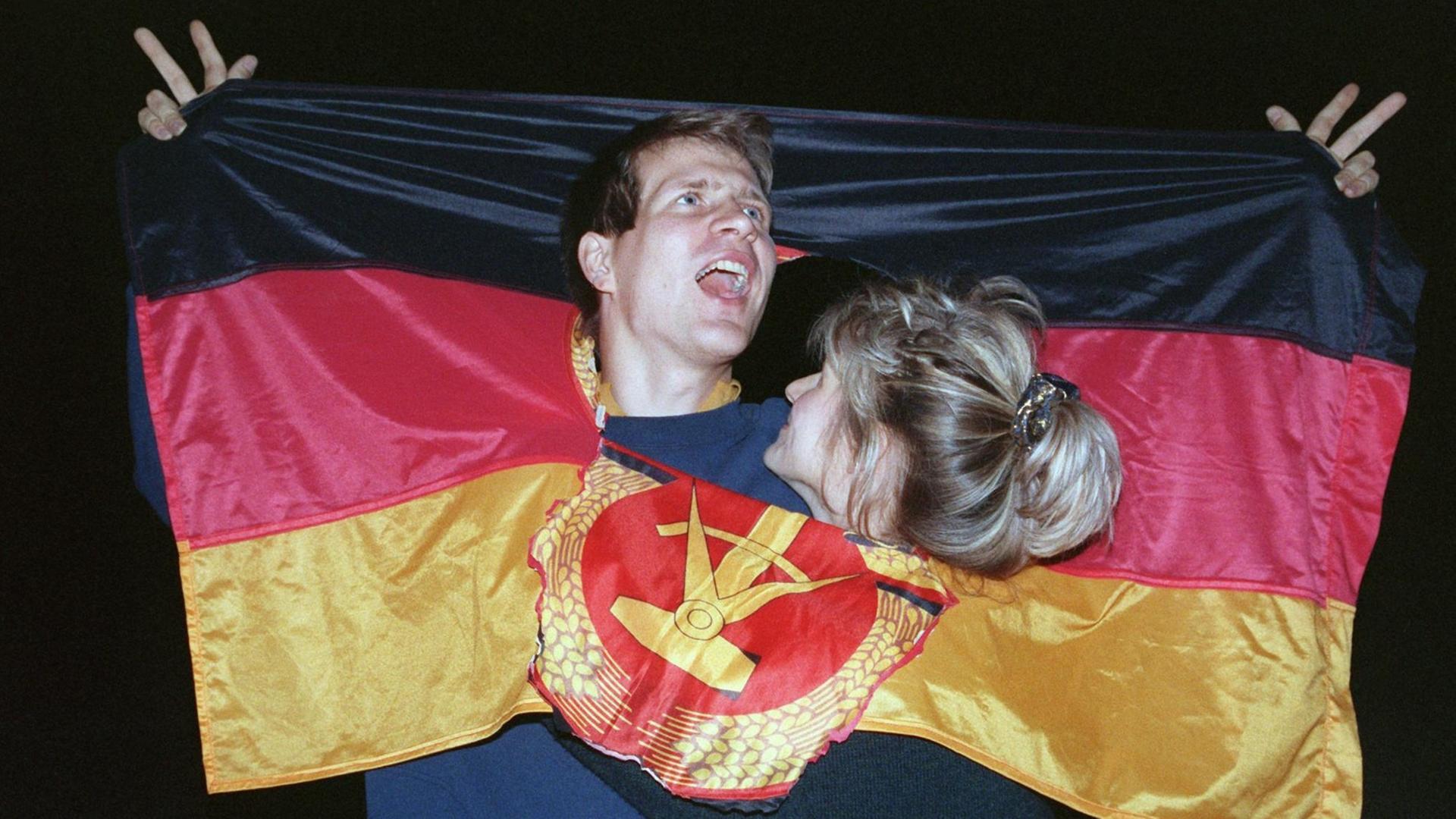 Ein jubelndes Paar hat sich in Berlin, am Tag der Deutschen Einheit in einer DDR-Fahne Platz geschaffen, wo früher das Emblem Hammer und Sichel war – und feiert den historischen Anlass. Ein jubelndes Paar hat sich in Berlin, am Tag der Deutschen Einheit in einer DDR-Fahne Platz geschaffen, wo früher das Emblem Hammer und Sichel war – und feiert den historischen Anlass.