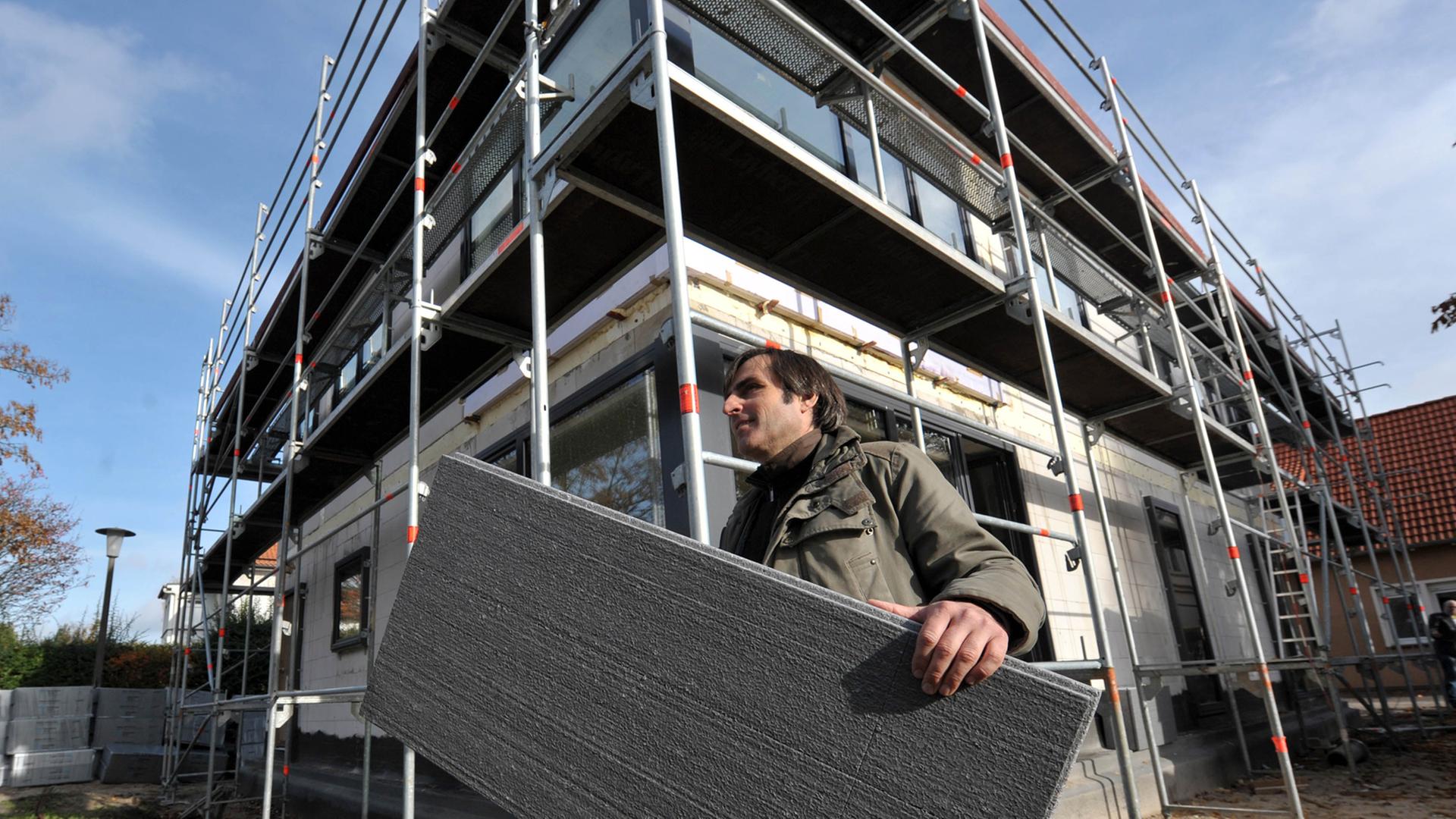 Ein Mann trägt vor dem Rohbau eines mehrstöckigen Hauses eine Wärmedämmplatte unter dem Arm. Ein Mann trägt vor dem Rohbau eines mehrstöckigen Hauses eine Wärmedämmplatte unter dem Arm.